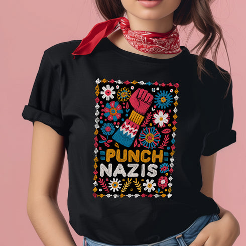 Black t-shirt featuring an embroidered-style design with the phrase "PUNCH NAZIS" in bold, colorful lettering. Above the text, a raised red fist is illustrated, symbolizing resistance, and is surrounded by a vibrant folk-art-inspired floral border with bright flowers, leaves, and geometric patterns in red, yellow, blue, and white.